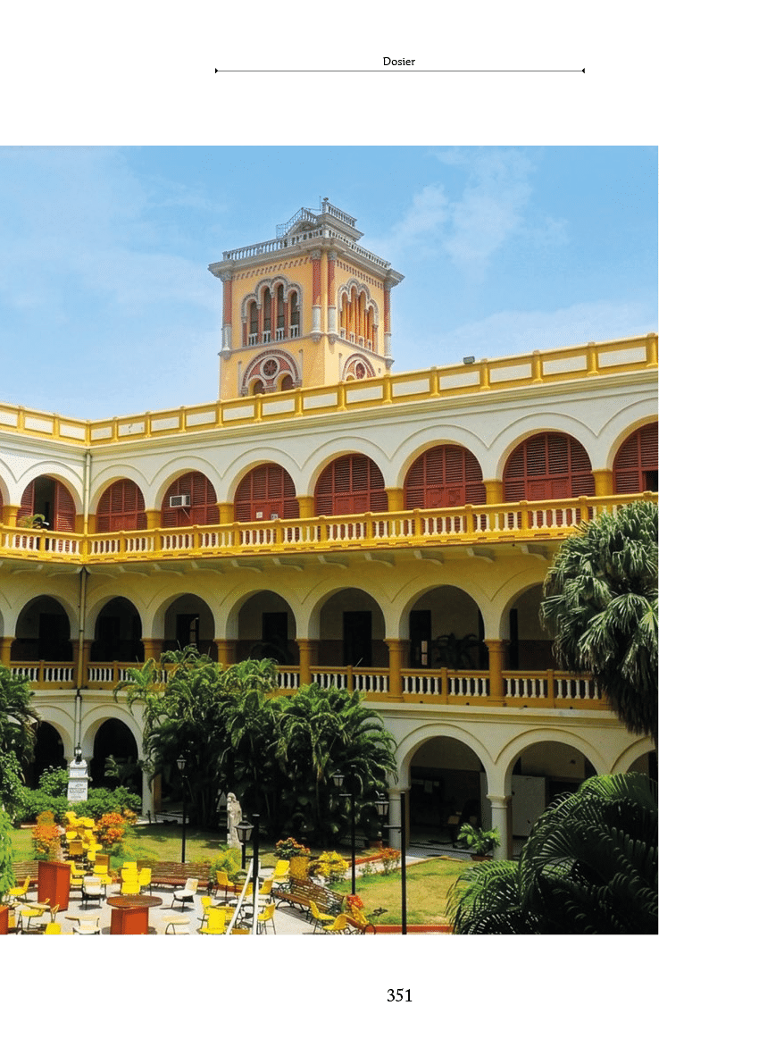 Edificio hist rico con arcos y balcones amarillos alrededor de un patio interior con vegetaci n y una torre alta al fondo bajo un cielo despejado. Descripci n generada con IA